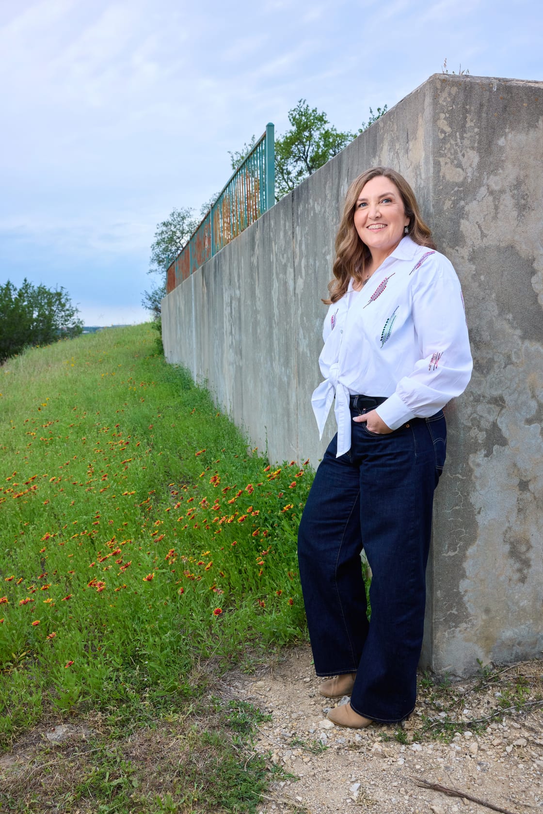 Keri Waters leaning against a concrete catchment wall along a path of Indian-blanket wildflowers in the Texas Hill Country.